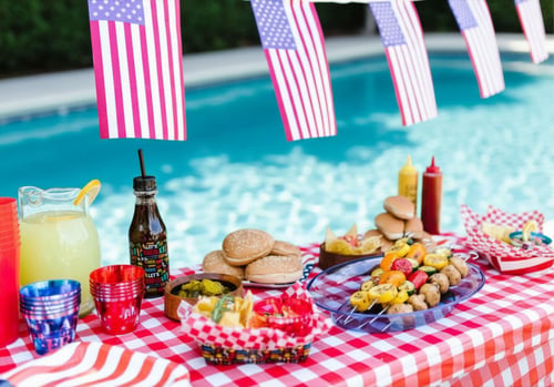 4th of July pool party food spread with lemonade, kabobs, and burgers on a red checkered tablecloth beside a backyard pool