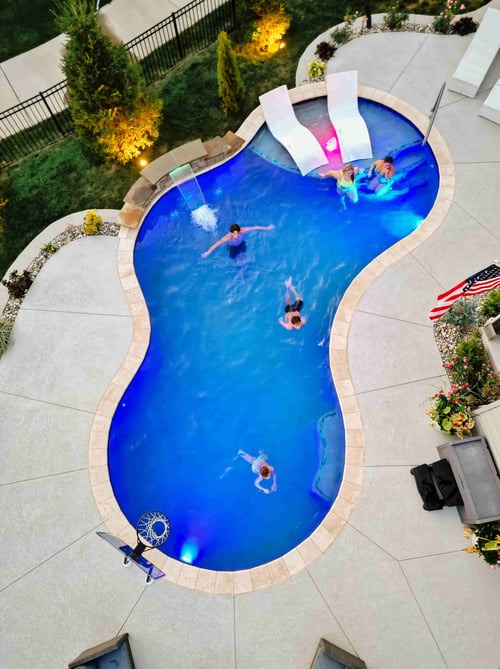 Aerial view of a freeform backyard swimming pool with several people swimming at dusk