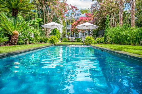 Long resort-style swimming pool surrounded by lush tropical landscaping, white umbrellas, flowering trees, and a house in the background.