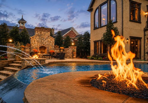Backyard pool with fire bowl and stone outdoor fireplace lit at dusk