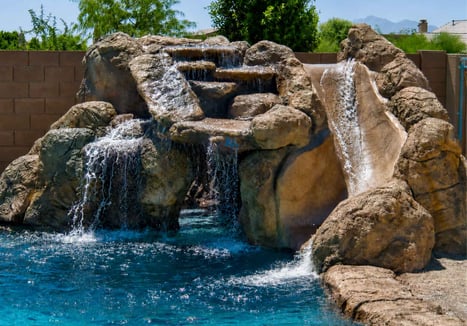 A decorative faux-rock waterfall and slide feature in a backyard swimming pool, with cascading water flowing into a bright blue pool on a sunny day.