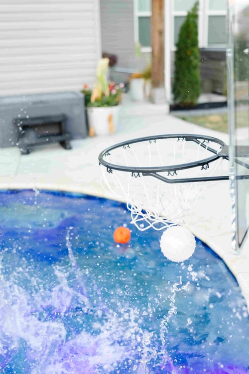 Poolside basketball hoop with ball mid-shot above splashing water in a backyard pool