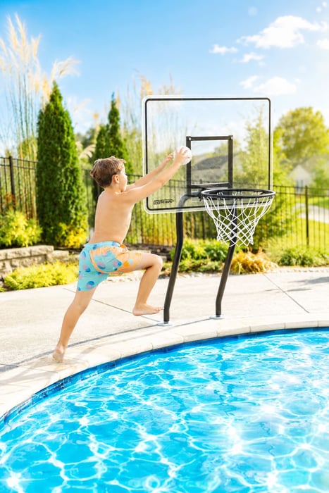 Young boy reaching up to dunk a basketball into a poolside basketball hoop in a backyard pool.
