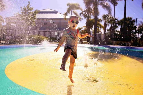 Young child in heart-shaped sunglasses joyfully running through water sprays at a colorful splash pad, with palm trees and sunlit sky in the background.