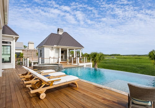 Infinity pool and hot tub on a wooden deck overlooking a Lowcountry marsh, with a white poolside cabana and teak lounge chairs in the foreground.