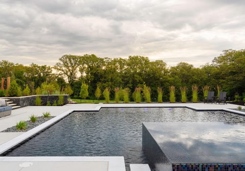 Finished inground concrete pool with white deck, modern design, lounge chairs, and green landscaping.
