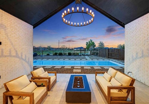 Covered outdoor living space with white brick walls, a round chandelier, and teak furniture surrounding a rectangular fire pit table, looking out to a lap pool and stepping stone pathway at dusk