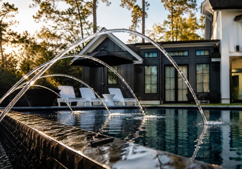 Multiple deck jets shooting arcing streams over a dark-tiled contemporary pool at golden hour