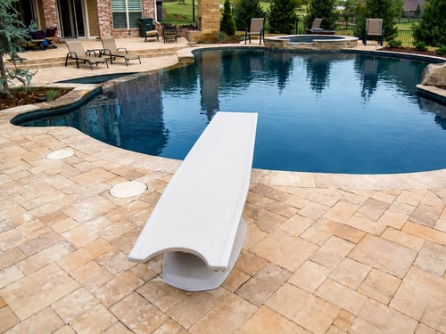 White diving board on a travertine pool deck next to a Blue Haven inground pool and spa