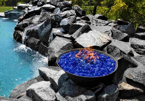 fire bowl filled with blue iridescent fire glass set into a large natural rock waterfall beside a pool