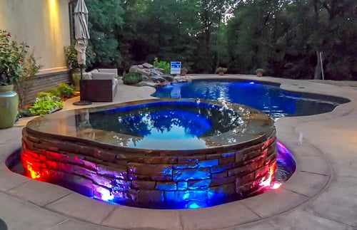 Organic-shaped rim-flow spa with multi-color LED lighting in a shaded backyard