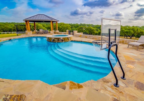 Freeform inground swimming pool with a sun shelf, poolside basketball hoop, raised circular spa with water features, and a wood-post gazebo, surrounded by a flagstone deck and wooded hill country views