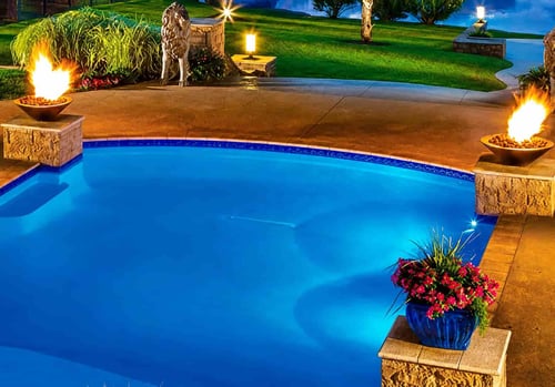 freeform backyard pool at night with two copper fire bowls on stone pedestals and landscape lighting