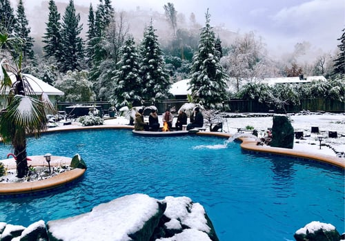 Group of friends sitting around a fire pit on a pool island surrounded by a heated swimming pool in the snow