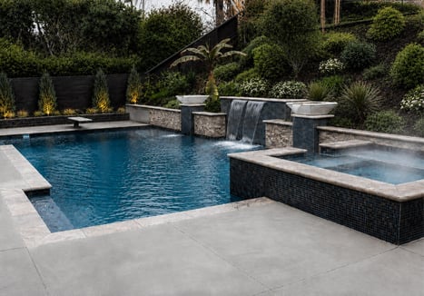 Inground pool and spa with stone waterfall feature, mosaic tile finish, and concrete pool deck surrounded by lush landscaping