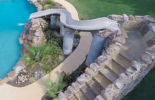 Elevated pool slide supported by columns with stone steps leading up beside landscaped pool