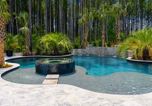 Luxury backyard swimming pool with turquoise water, circular raised seating platform in the center, light gray concrete decking, stone walls, and lush tropical landscaping including palm trees and tall pine forest backdrop