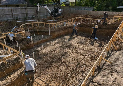 Construction site showing an excavated swimming pool foundation with metal rebar reinforcement, wooden framing forms, multiple workers, and a mini excavator in a residential backyard with wooden fencing