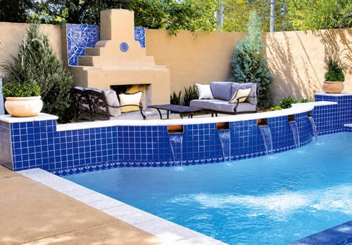 Pool patio with built-in outdoor fireplace, curved blue tile raised wall with waterfall scuppers, and cushioned outdoor sofas