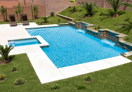 Geometric pool and spa with white deck on the house side only, bordered by a landscaped grass slope with no deck on the far side
