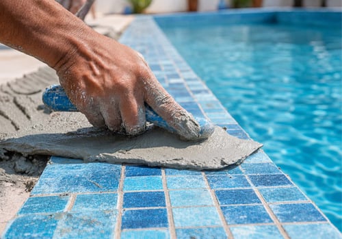 Close-up of a worker's hand covered in wet gray cement, applying sealant or grout to the blue tiled edge of a swimming pool during repair or maintenance work