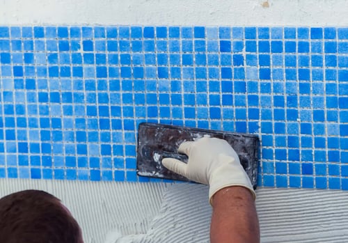 Worker applying pool tile finish with a trowel on blue mosaic tiles during inground pool construction.