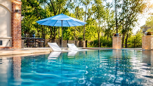 Blue umbrella set into a tanning ledge in a backyard swimming pool across the pool with lounge chairs