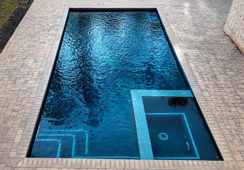 Aerial view of a rectangular inground swimming pool with deep blue water, submerged steps and a spa section outlined in light blue mosaic tile, surrounded by a gray paver deck