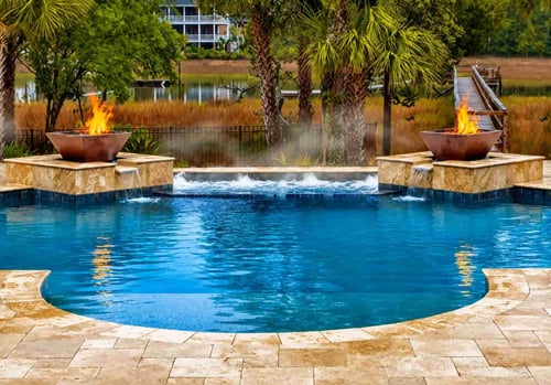 two square copper fire bowls on travertine pedestals flanking a freeform pool with spa spillover and waterfront background